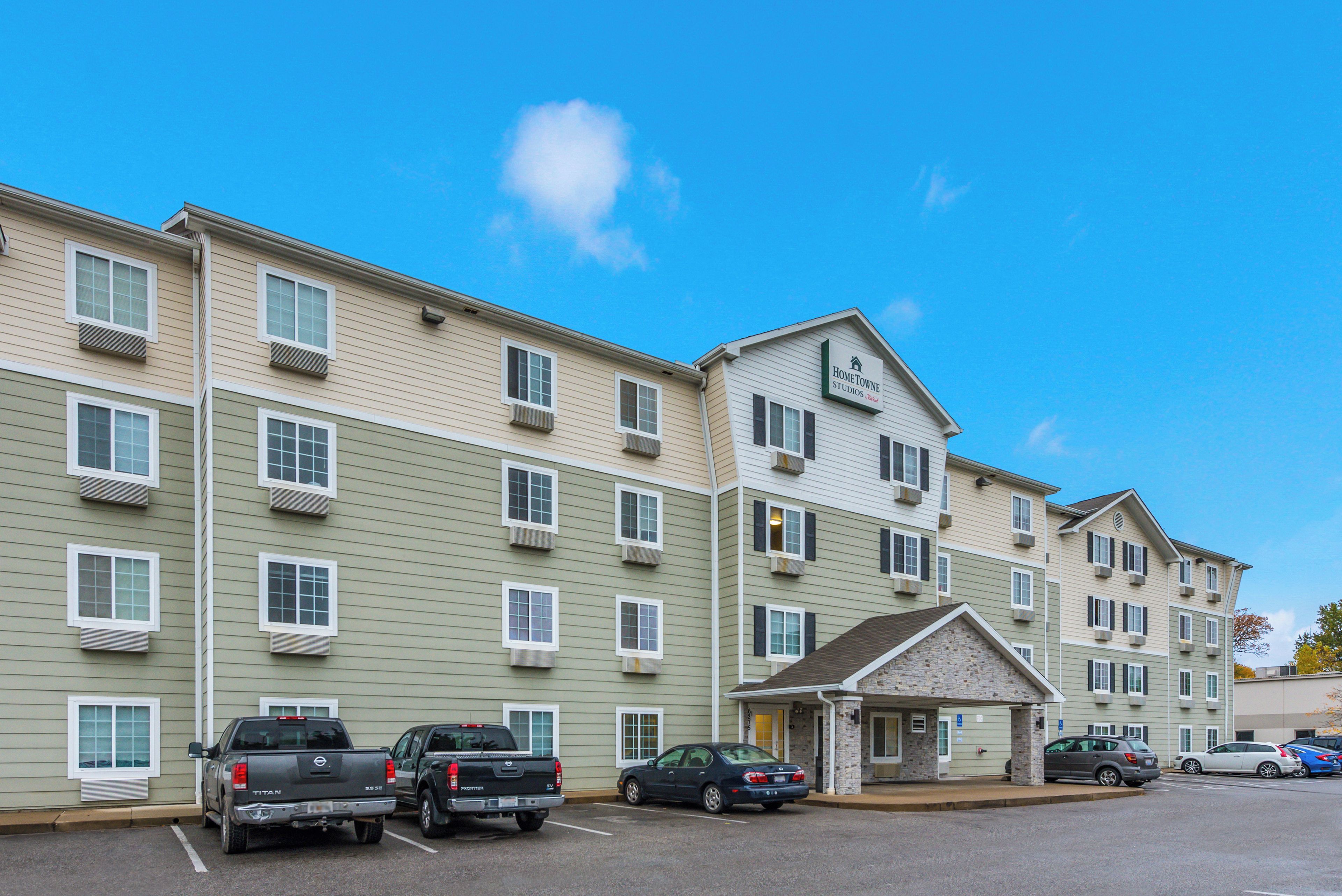 A multi-story hotel with a central entrance, parking lot, and several vehicles parked in front.