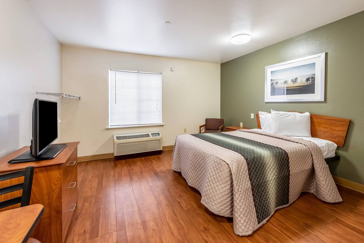 A bedroom with a bed, a desk with a computer, a chair, a window with blinds, and a picture on the wall.