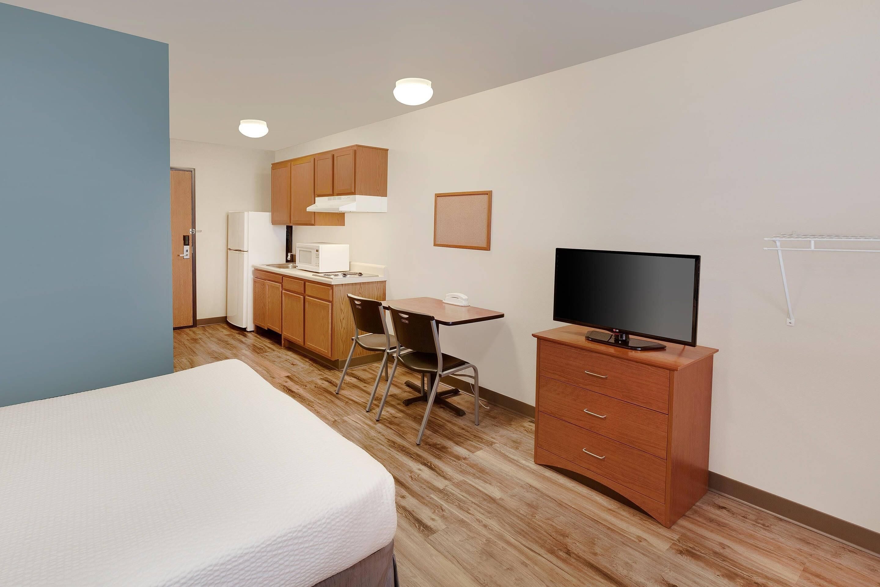A compact hotel room with a bed, kitchenette, TV, and a small desk.