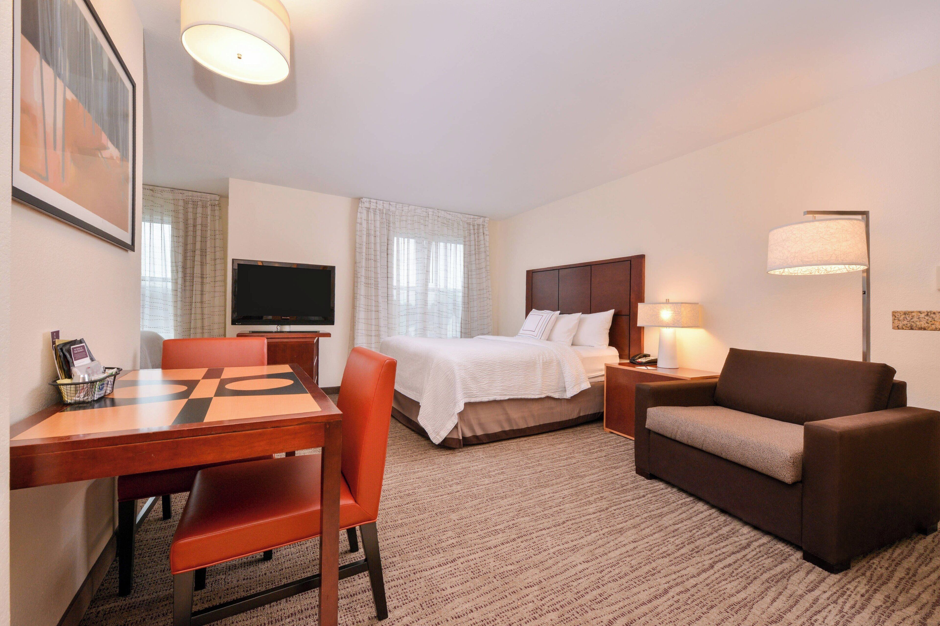 A hotel room with a bed, a sofa, a TV, a desk with a chair, and a lamp.