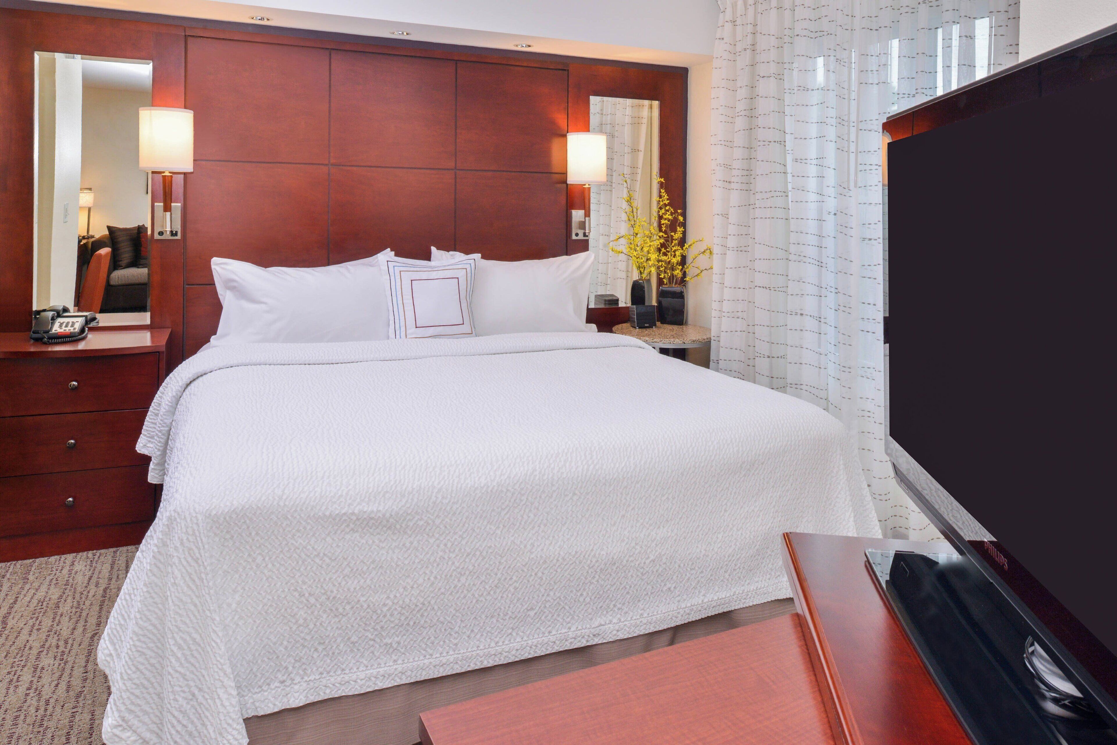 Habitación de hotel con una cama grande, cabecera de madera, mesita de noche con lámpara, televisor y espejo.