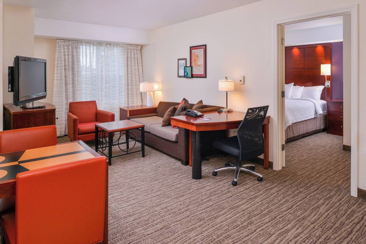 Quarto de hotel com cama, escrivaninha, cadeira, sofá, televisão e luminária.
