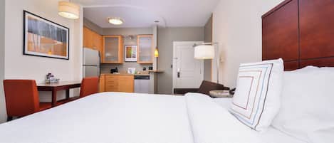 A hotel room with a large bed, a desk, a chair, a small kitchen area, and a dining table.
