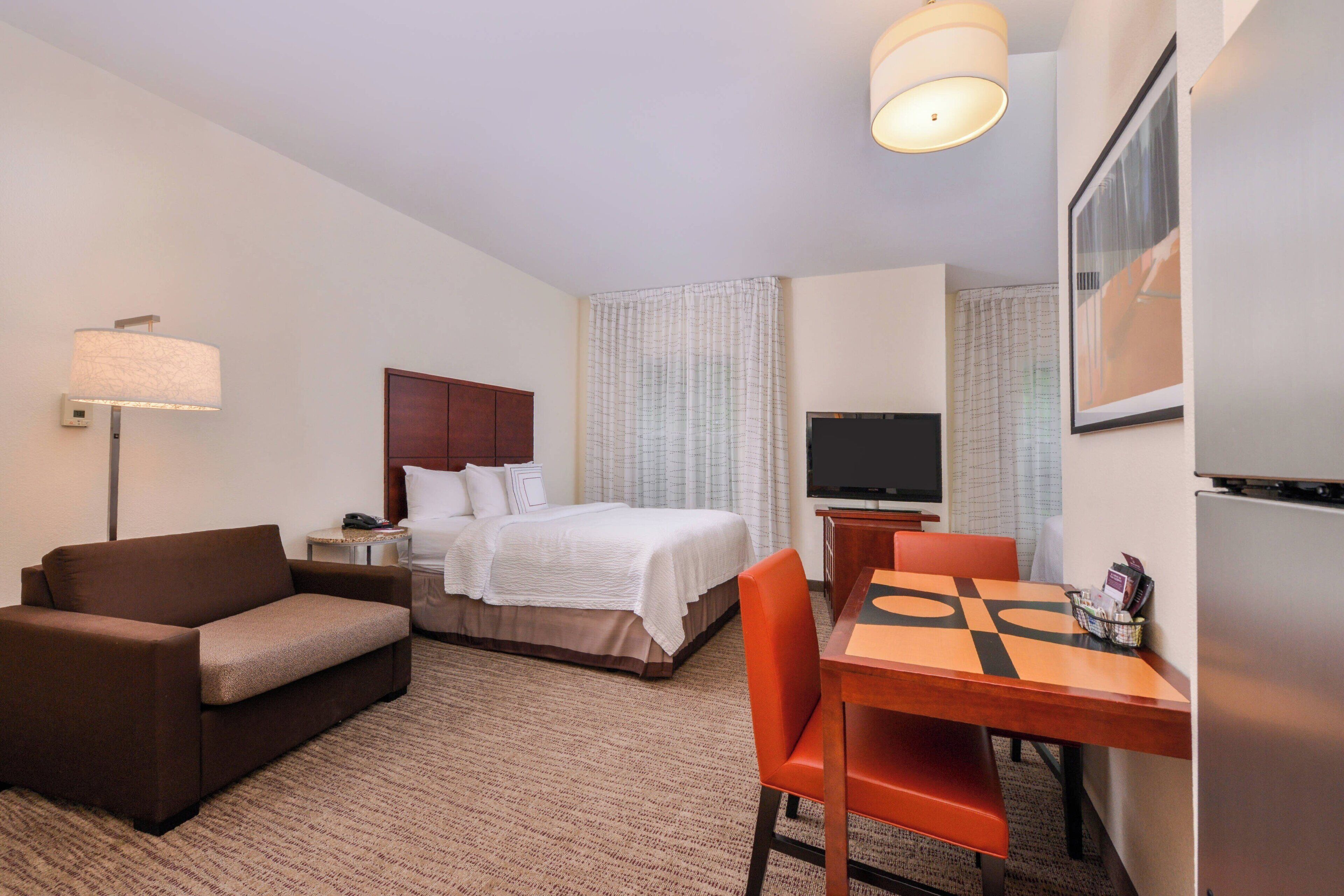 A hotel room with a bed, a desk with a chair, a sofa, a lamp, a TV, and a refrigerator.