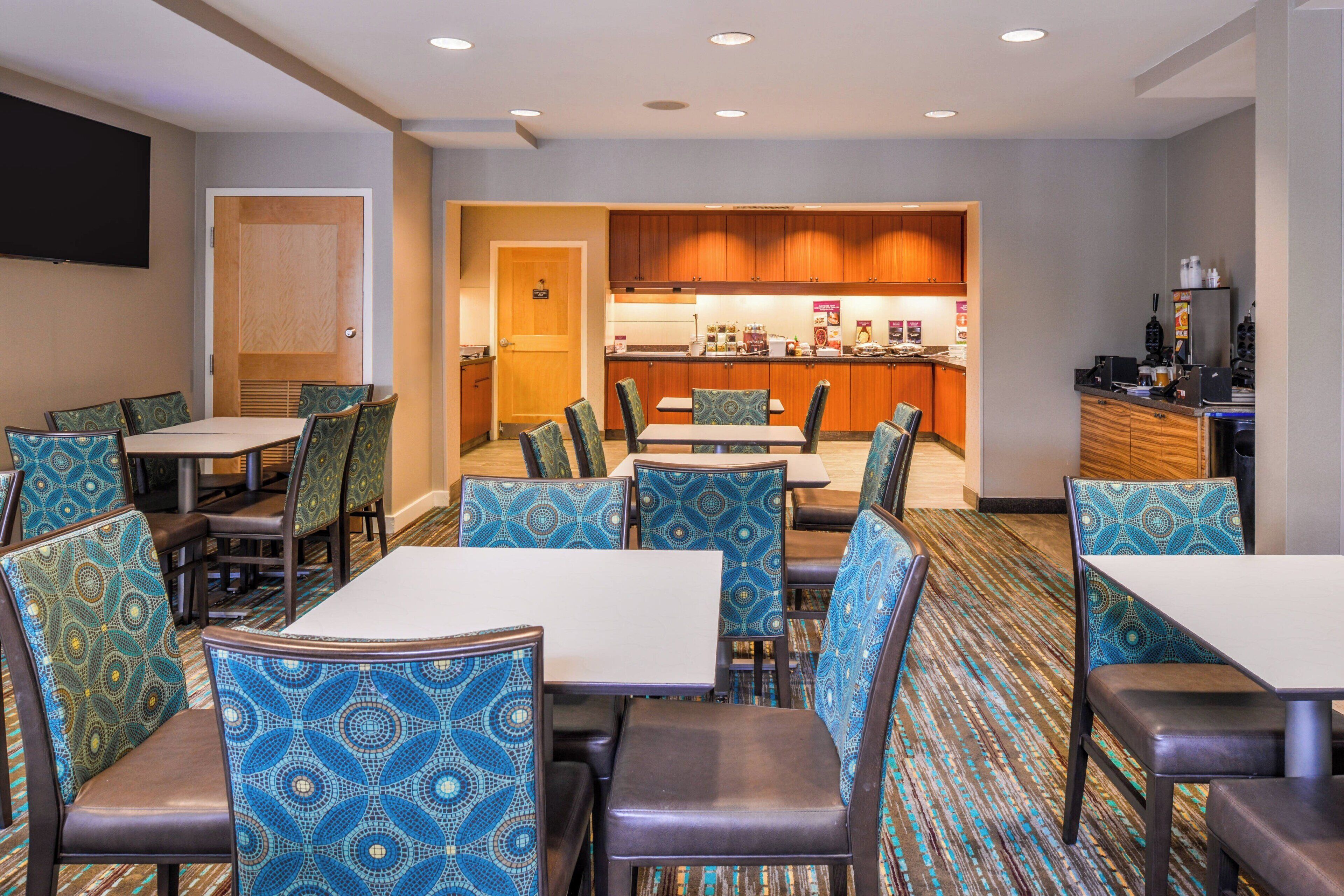 A hotel dining area with patterned chairs and tables, a kitchen in the background, and a flat-screen TV mounted on the wall.