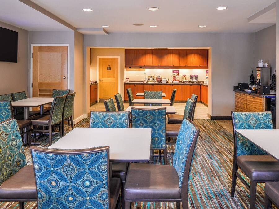 A hotel dining area with patterned chairs and tables, a kitchen in the background, and a flat-screen TV mounted on the wall.
