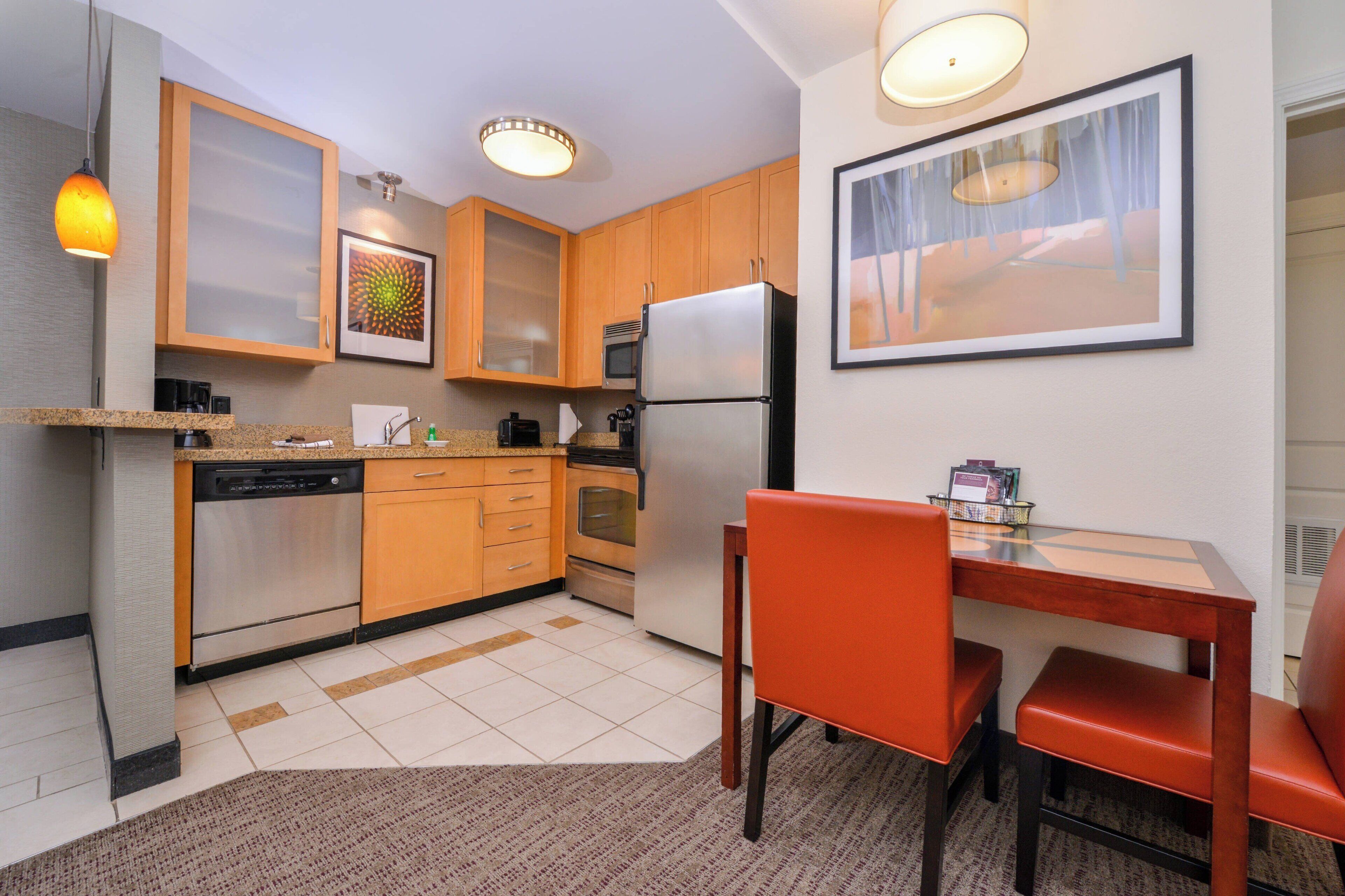 Una cocina compacta con armarios de madera, un refrigerador de acero inoxidable y una mesa de comedor con sillas anaranjadas.