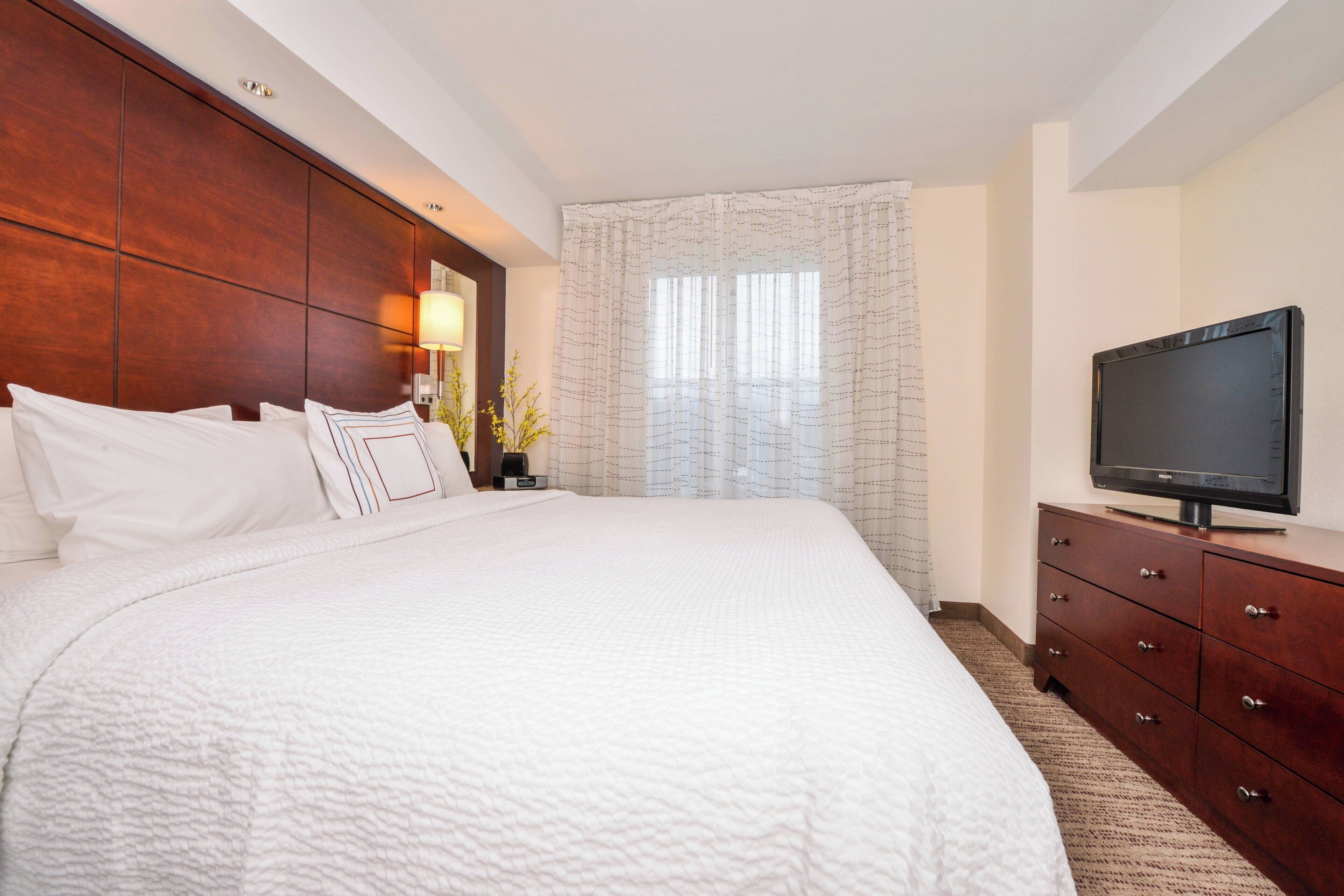 A hotel room with a large bed, a wooden headboard, a television on a dresser, and a window with sheer curtains.
