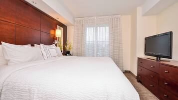 A hotel room with a large bed, a wooden headboard, a television on a dresser, and a window with sheer curtains.