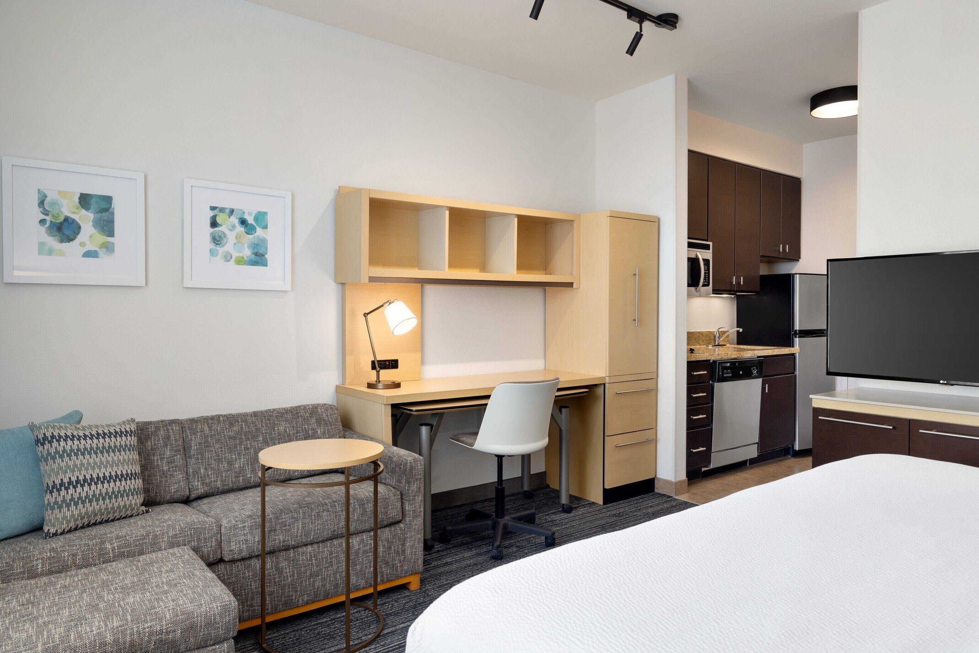 A hotel room with a bed, a sofa, a desk with a chair, a small table, a TV, a kitchen area with cabinets and appliances, and two framed artworks on the wall.