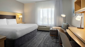 Ein Hotelzimmer mit Bett, Schreibtisch, Stuhl, einem kleinen Tisch, einer Klimaanlage, einer Lampe und einem fenster mit Vorhängen.