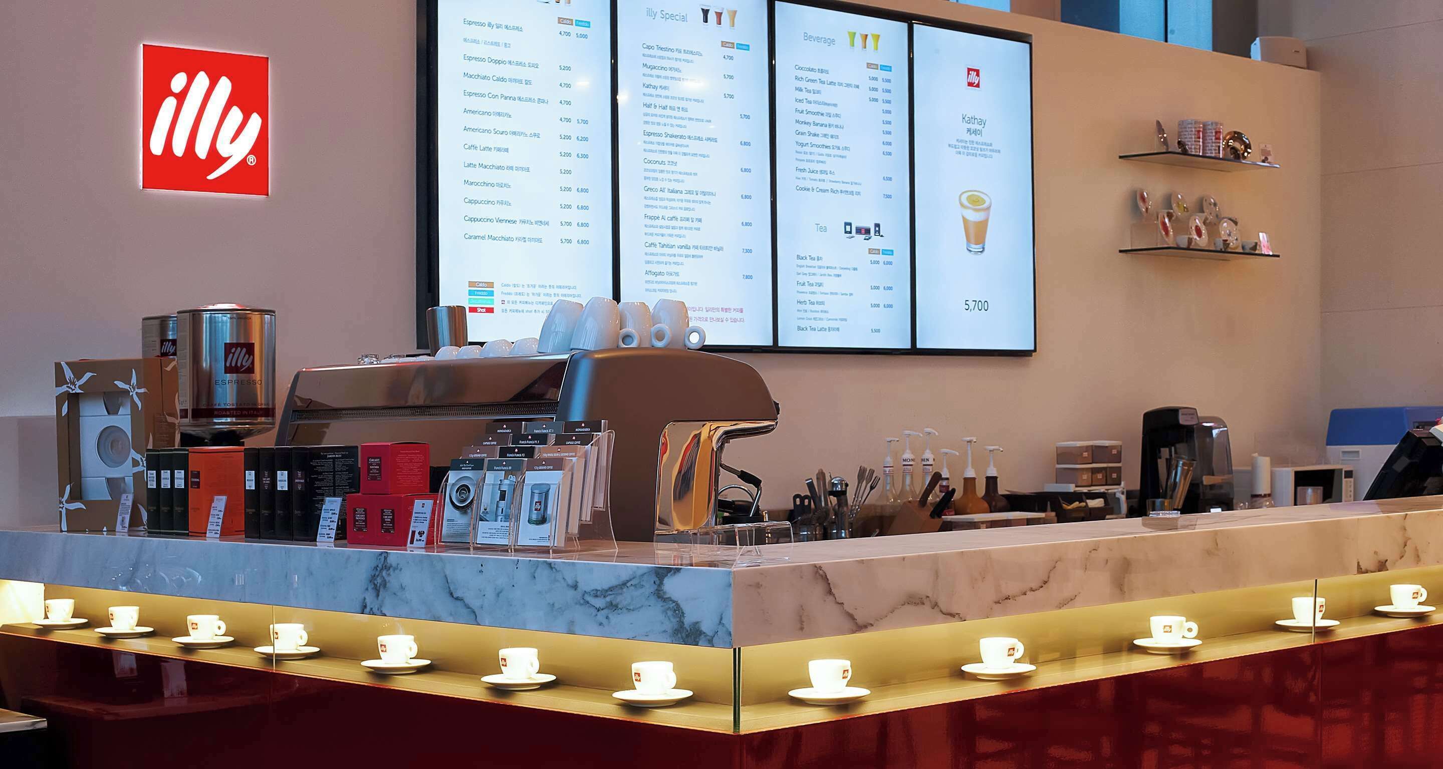 A coffee shop counter with a menu board, coffee machines, and a display of cups.
