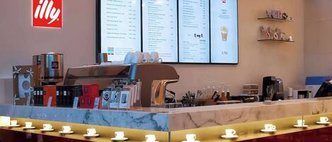 A coffee shop counter with a menu board, coffee machines, and a display of cups.