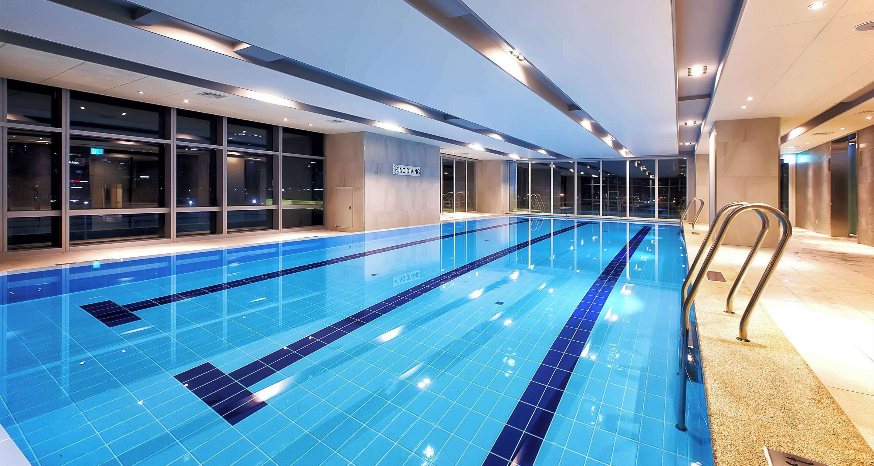 Una piscina interior con baldosas azules cristalinas, un cartel que indica 'No Diving' y un diseño arquitectónico moderno con ventanales amplios y suelo de baldosas.