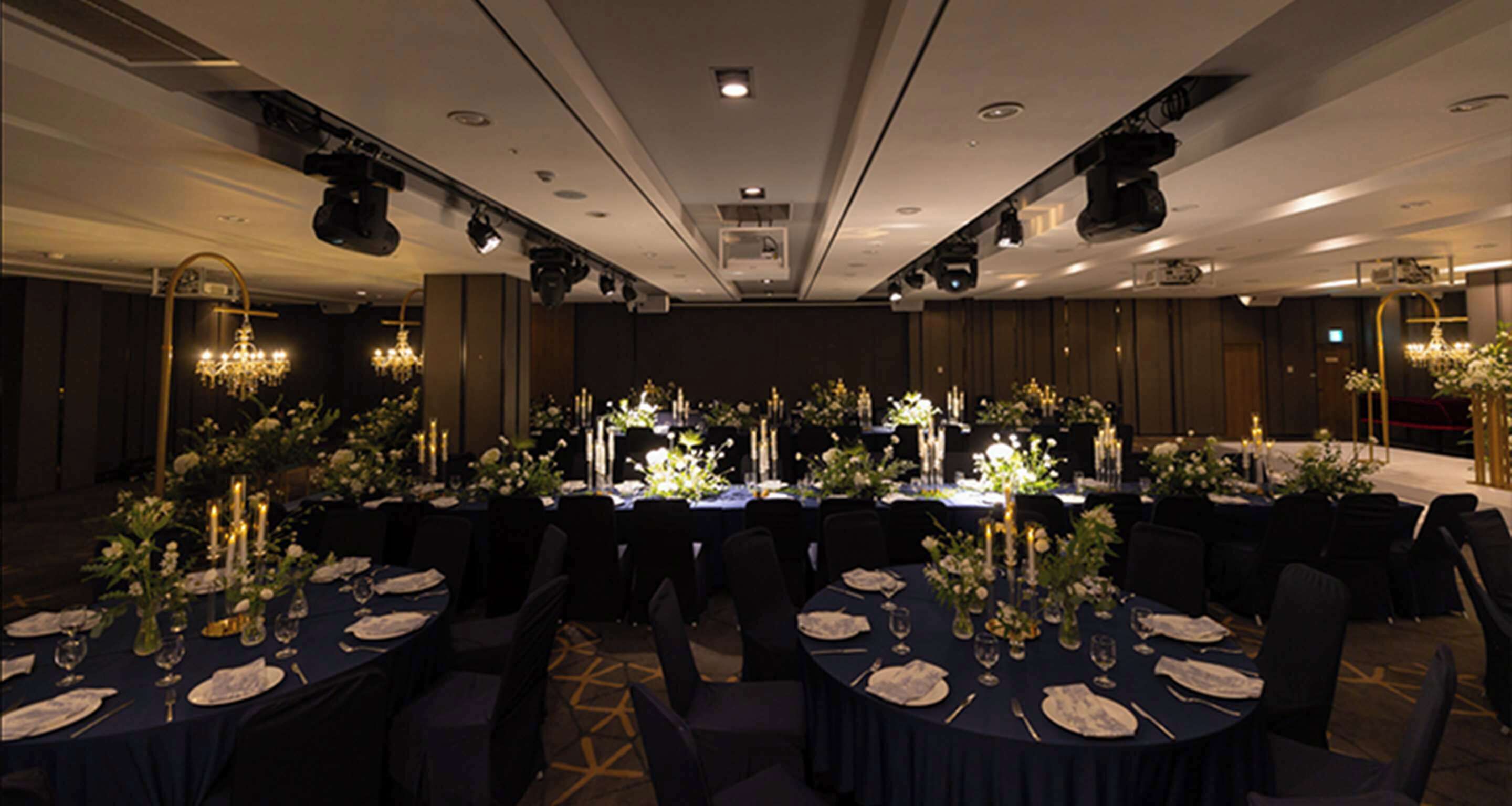 Une salle de banquet avec des tables rondes dressées pour le repas, chacune étant recouverte d’une nappe blanche et agrémentée de bougies et de compositions florales.