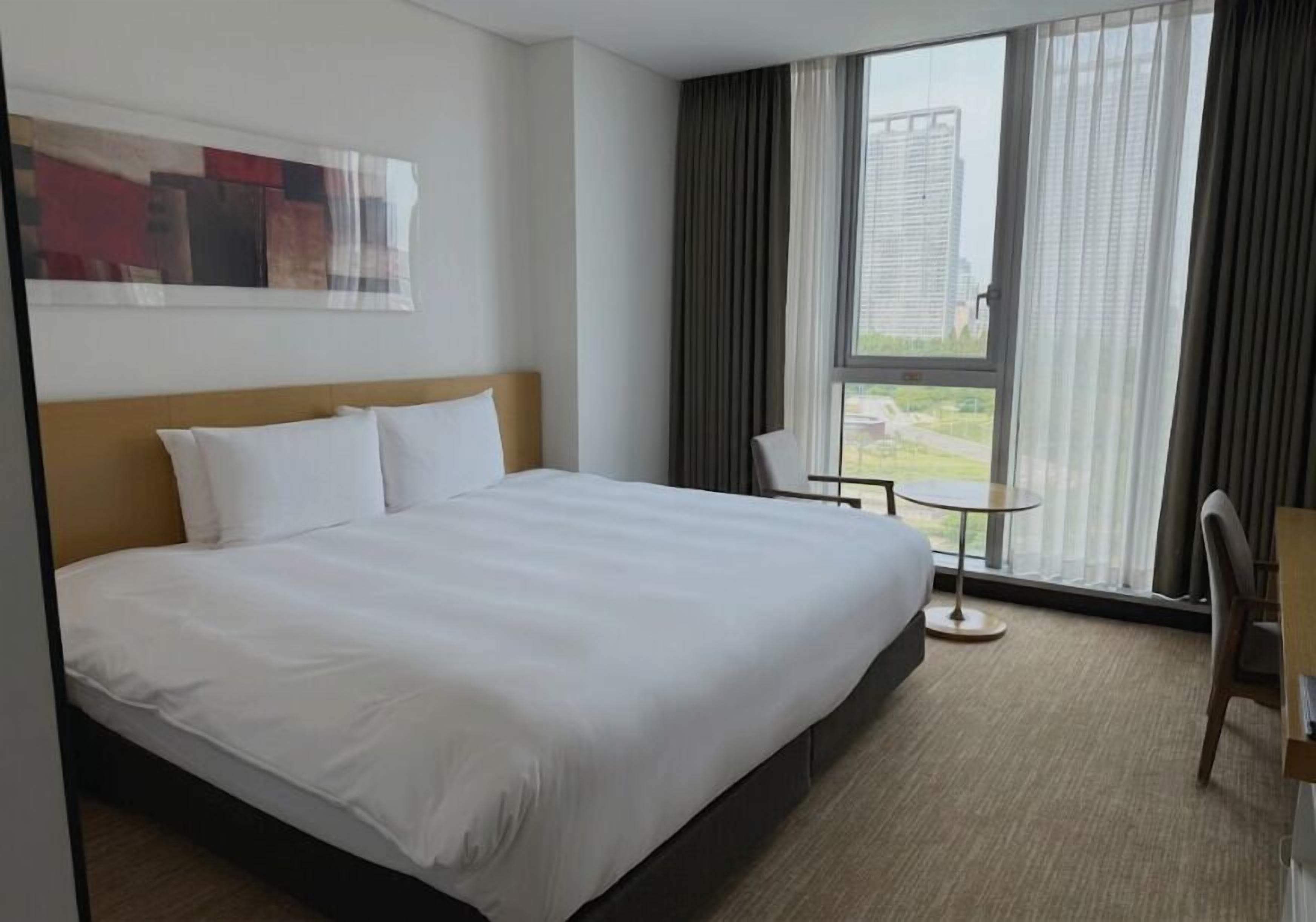 A hotel room with a large bed, a small table, a chair, and a view of buildings through the window.
