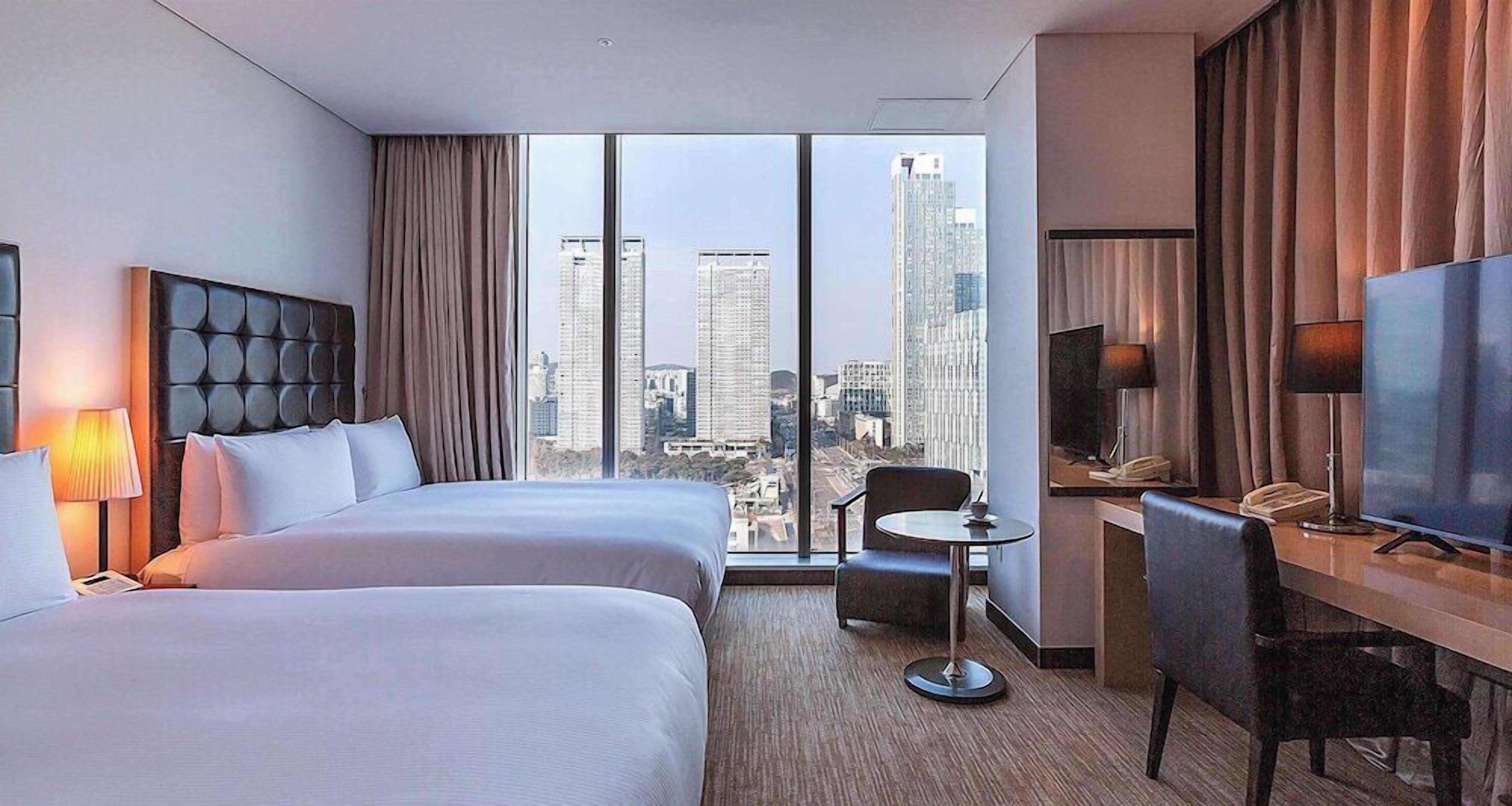 A hotel room with two beds, a desk, a chair, a TV, and a view of city buildings.