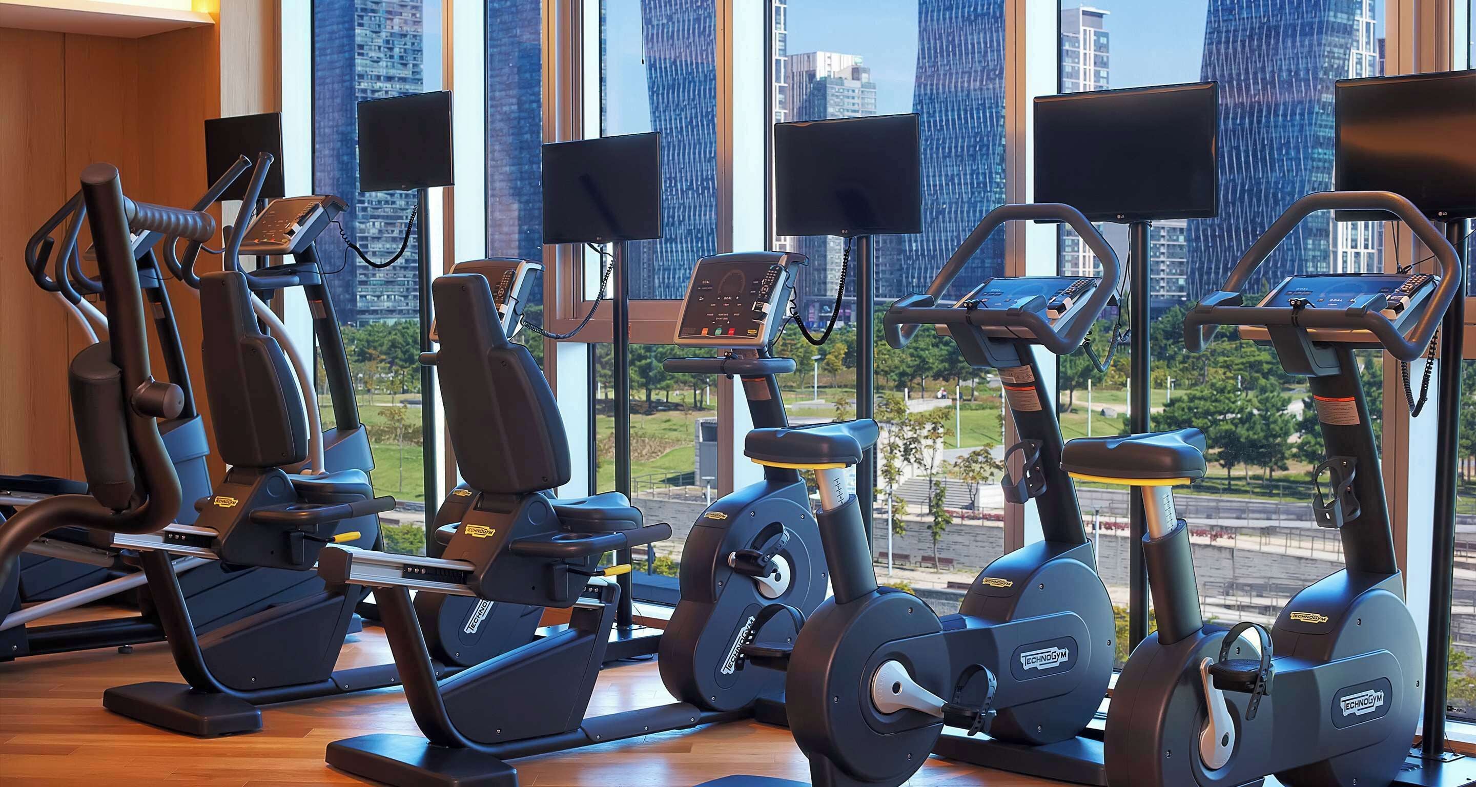 A gym with various exercise machines and large windows offering city views.