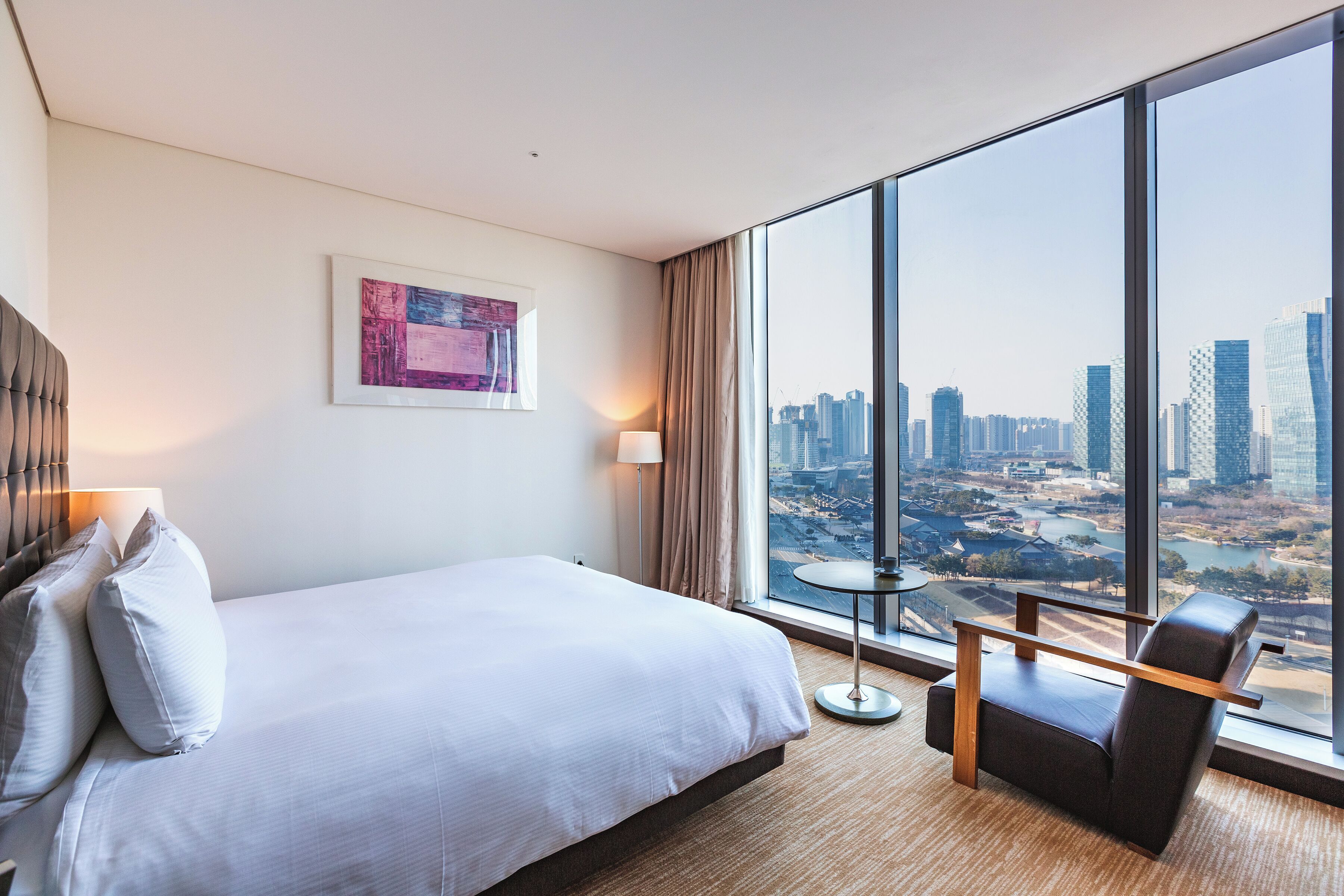 A hotel room with a large bed, a chair, a small table, a lamp, and a city view through large windows.