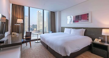 Habitación de hotel con una cama grande, un escritorio con teléfono, una silla, una mesita y vistas a la ciudad a través de amplias ventanas.