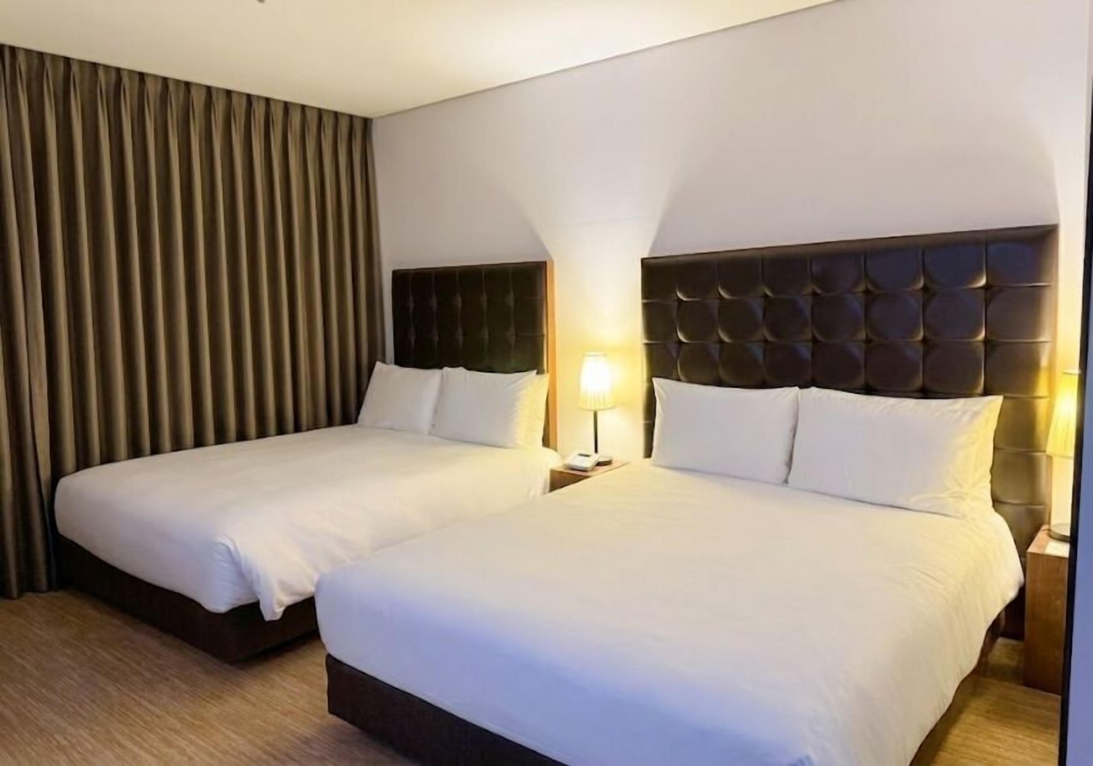 A hotel room with two beds, a dark headboard, a nightstand with a lamp, and a large window with curtains.