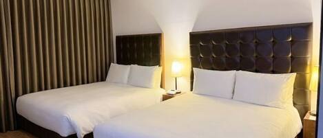 A hotel room with two beds, a dark headboard, a nightstand with a lamp, and a large window with curtains.