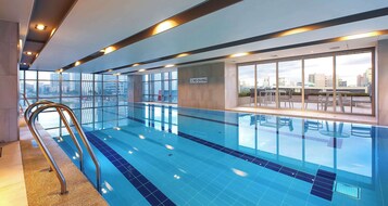 Una piscina interior con paredes de vidrio transparente, una escalera metálica y vista a la ciudad.
