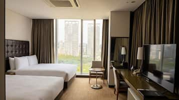 Habitación de hotel con dos camas, un escritorio con televisor, una silla y un ventanal con vista a la ciudad.