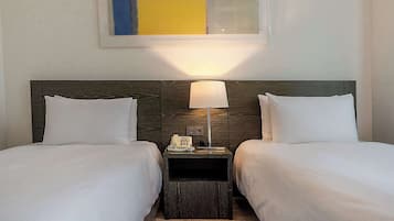 Una habitación de hotel con dos camas individuales, una mesita de noche con lámpara, un teléfono y una pequeña estantería.