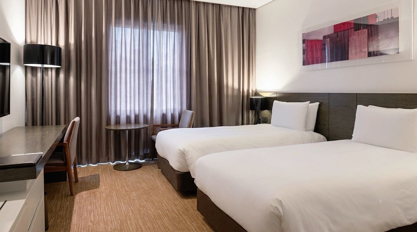 Habitación de hotel con dos camas, un escritorio, una silla, una mesa redonda y un ventanal con cortinas.