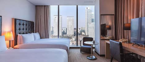 A hotel room with two beds, a desk, a chair, a TV, and a view of a cityscape.