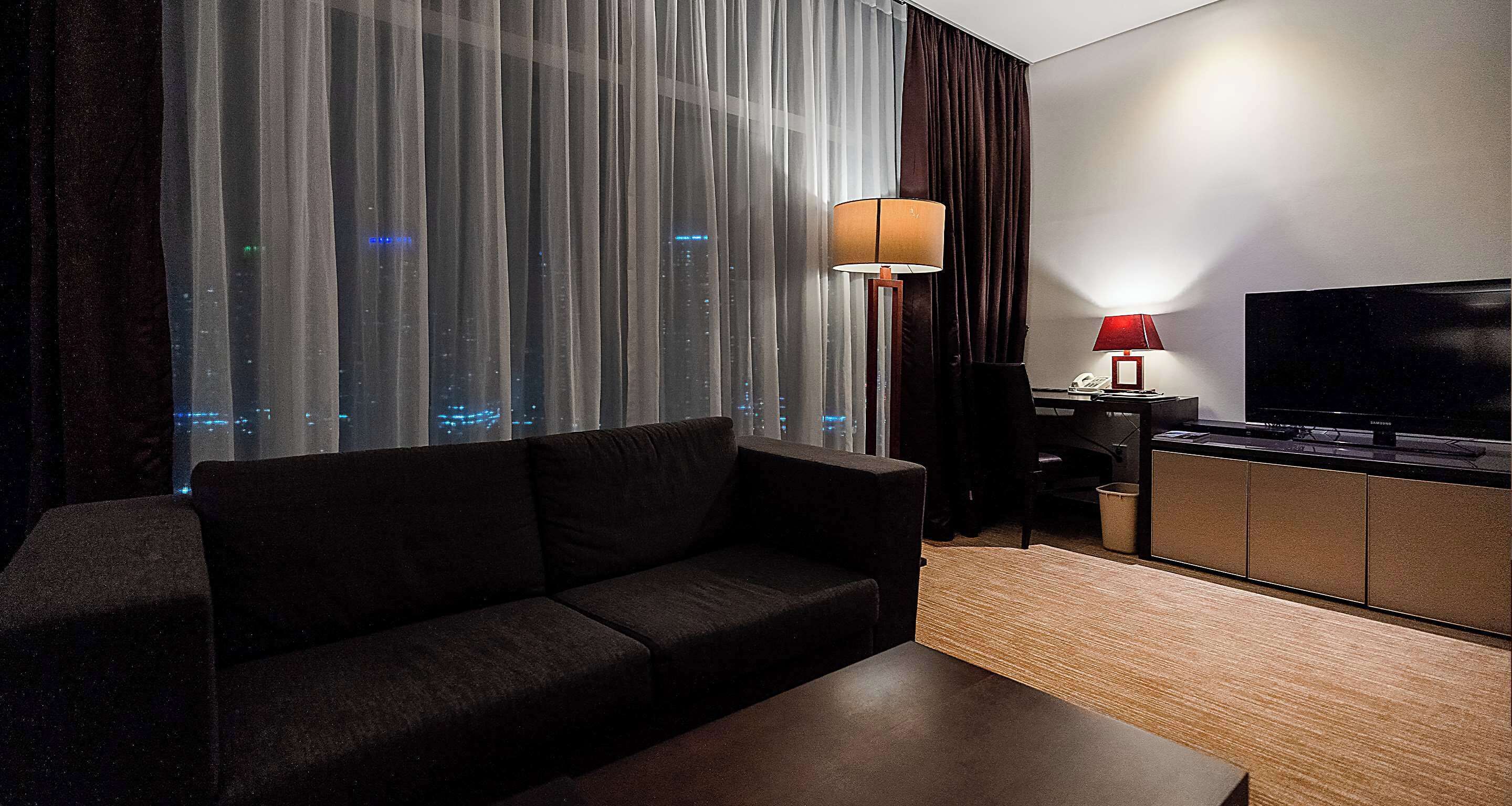 Habitación de hotel moderna con un sofá oscuro, una mesa de centro de madera, un televisor en un mueble y vistas a la ciudad a través de cortinas traslúcidas.