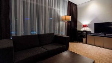 Habitación de hotel moderna con un sofá oscuro, una mesa de centro de madera, un televisor en un mueble y vistas a la ciudad a través de cortinas traslúcidas.