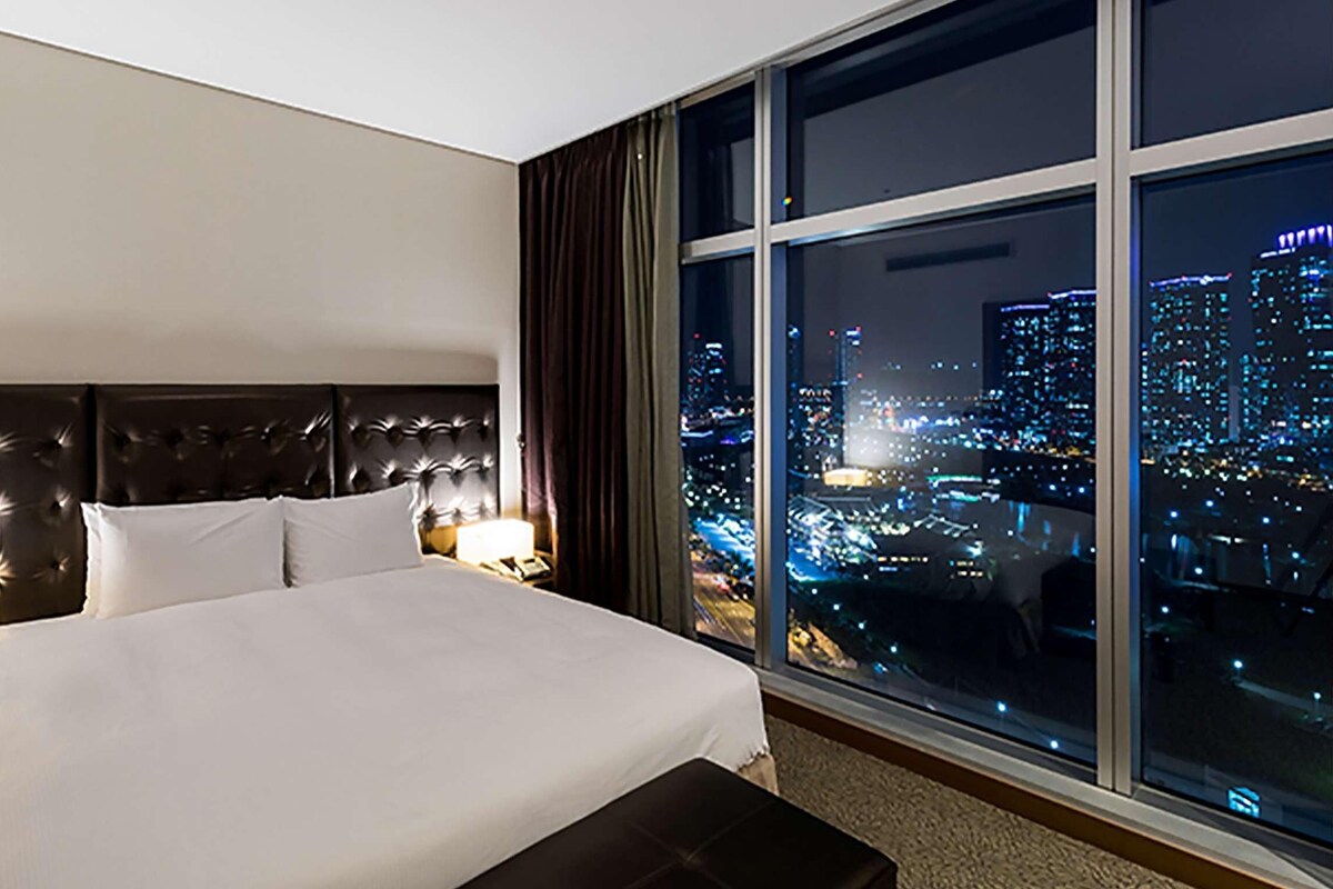 A hotel room with a large bed, a nightstand with a lamp, and a view of a cityscape at night.