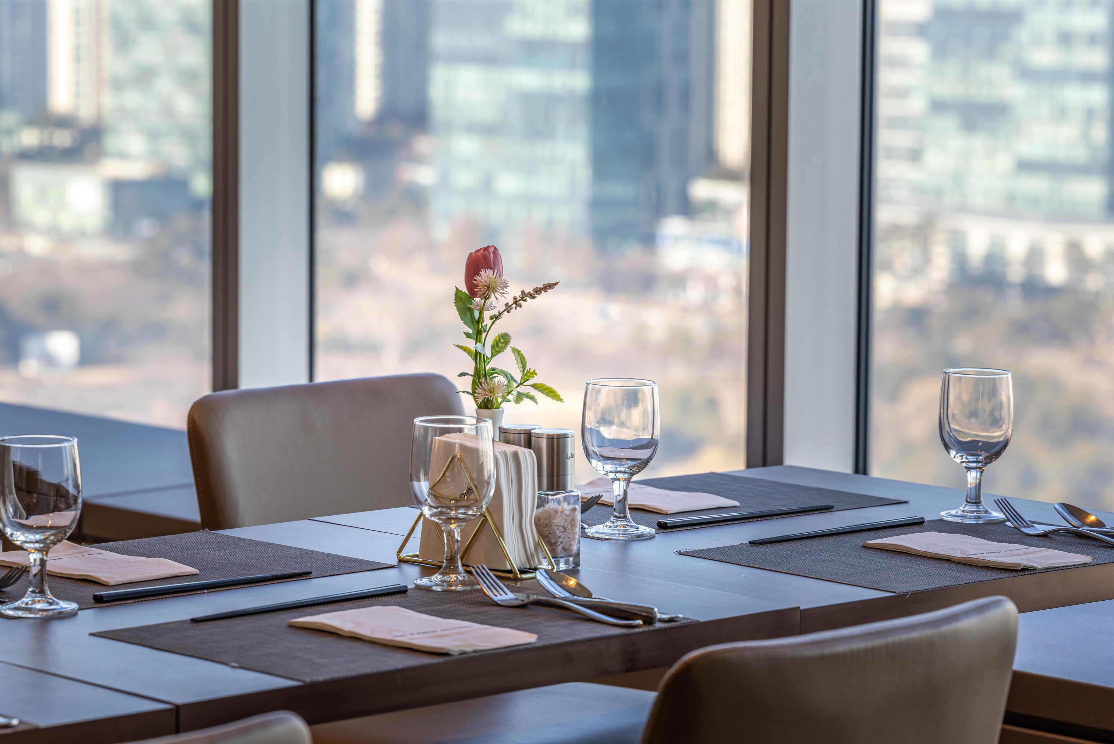 Una mesa de comedor lista con vasos, cubiertos y un florero con una flor, con vistas a un paisaje urbano a través de grandes ventanales.