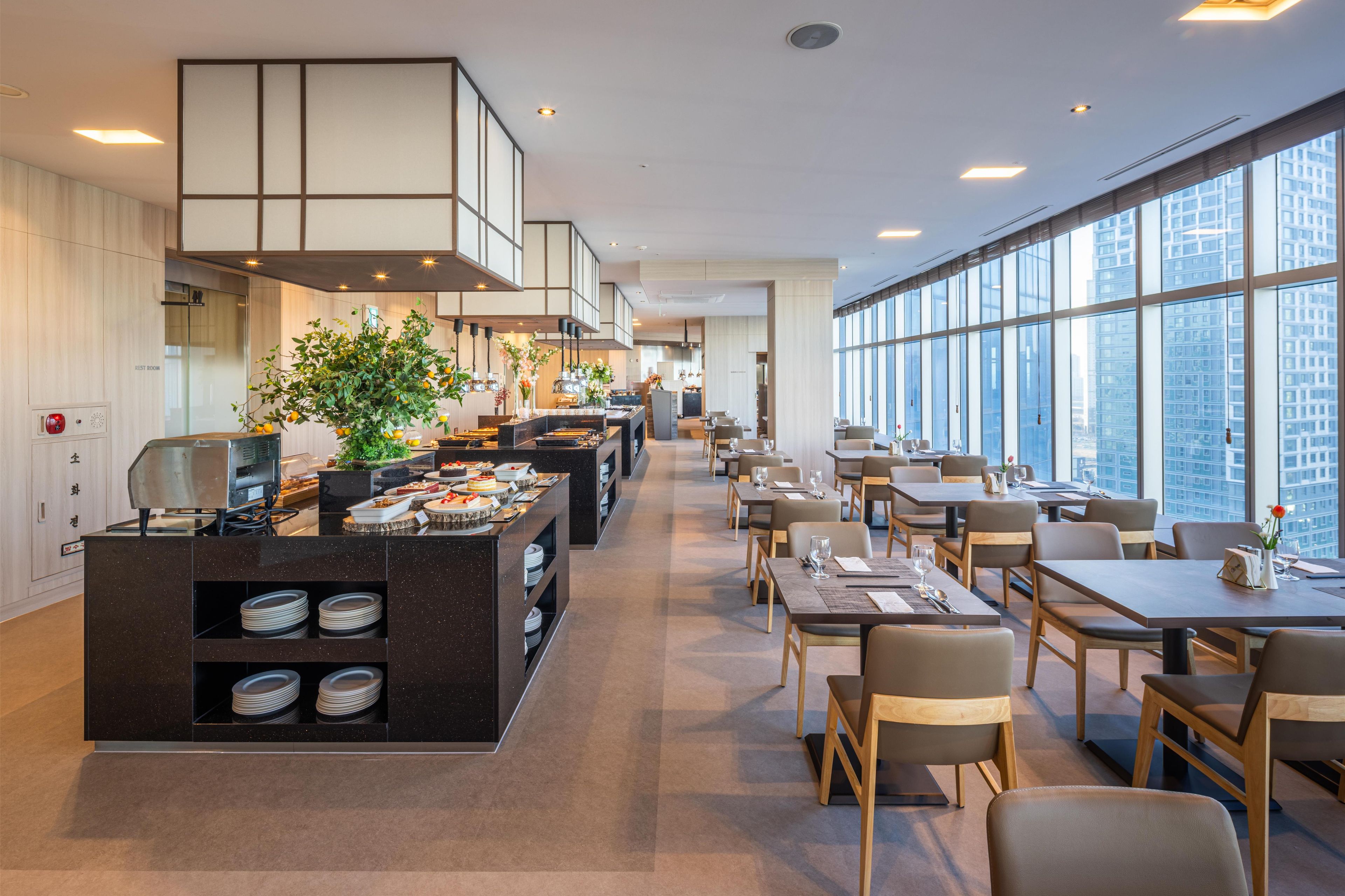 Un restaurant moderne doté d’un espace buffet, de tables et offrant une vue sur le paysage urbain grâce à de grandes fenêtres.