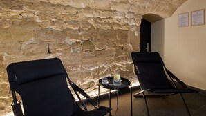 A room with stone walls, two reclining chairs, a small table with a glass and a bottle, and two informational posters on the wall. - Hotel Les Théâtres (Paris)