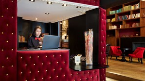 A reception area with a red velvet reception desk, a laptop, a tall vase with dried flowers, and a bookshelf filled with books. - Hotel Les Théâtres (Paris)