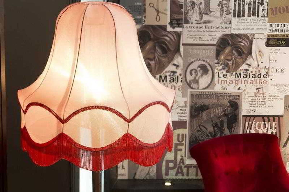 Eine Lampe mit gewelltem Schirm und rotem Fuß, vor einer Wand mit verschiedenen Postern und einem roten Stuhl platziert.