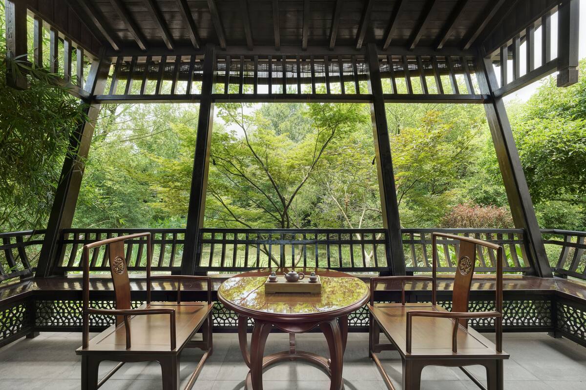 A traditional wooden pavilion with a round table and chairs, surrounded by a lattice railing, overlooking a lush green garden.