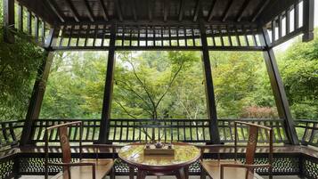 A traditional wooden pavilion with a round table and chairs, surrounded by a lattice railing, overlooking a lush green garden.