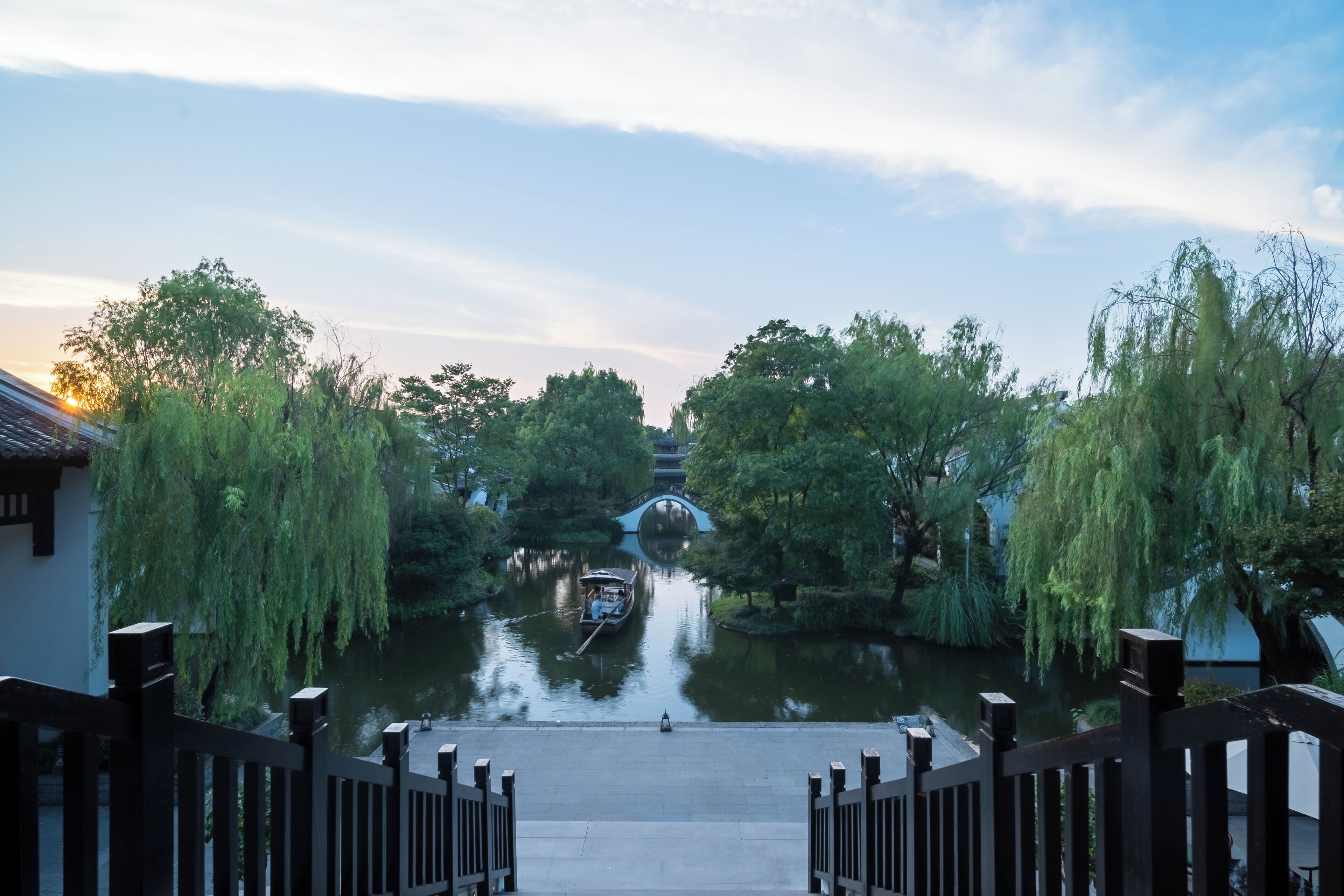 A riverside walkway with a traditional bridge, surrounded by trees and a calm water body.