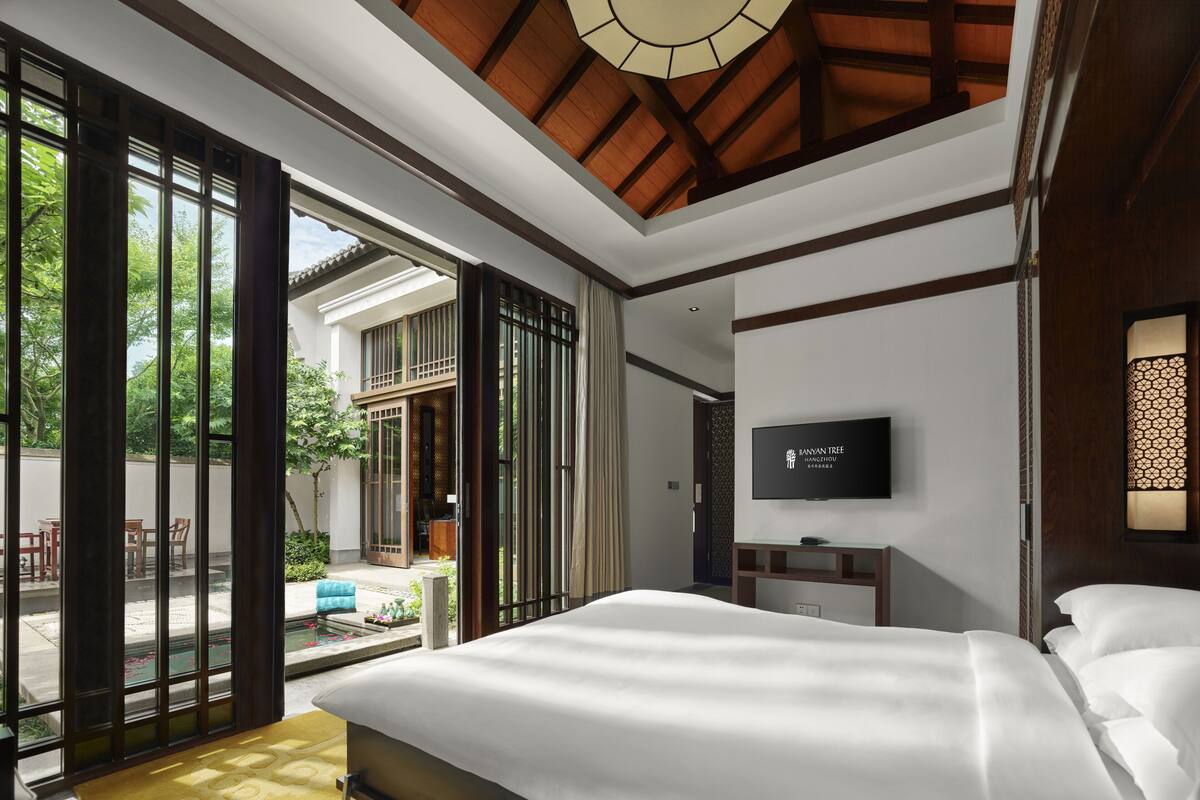 A modern hotel room with a large bed, a TV mounted on the wall, and a sliding glass door leading to an outdoor area with a pool.