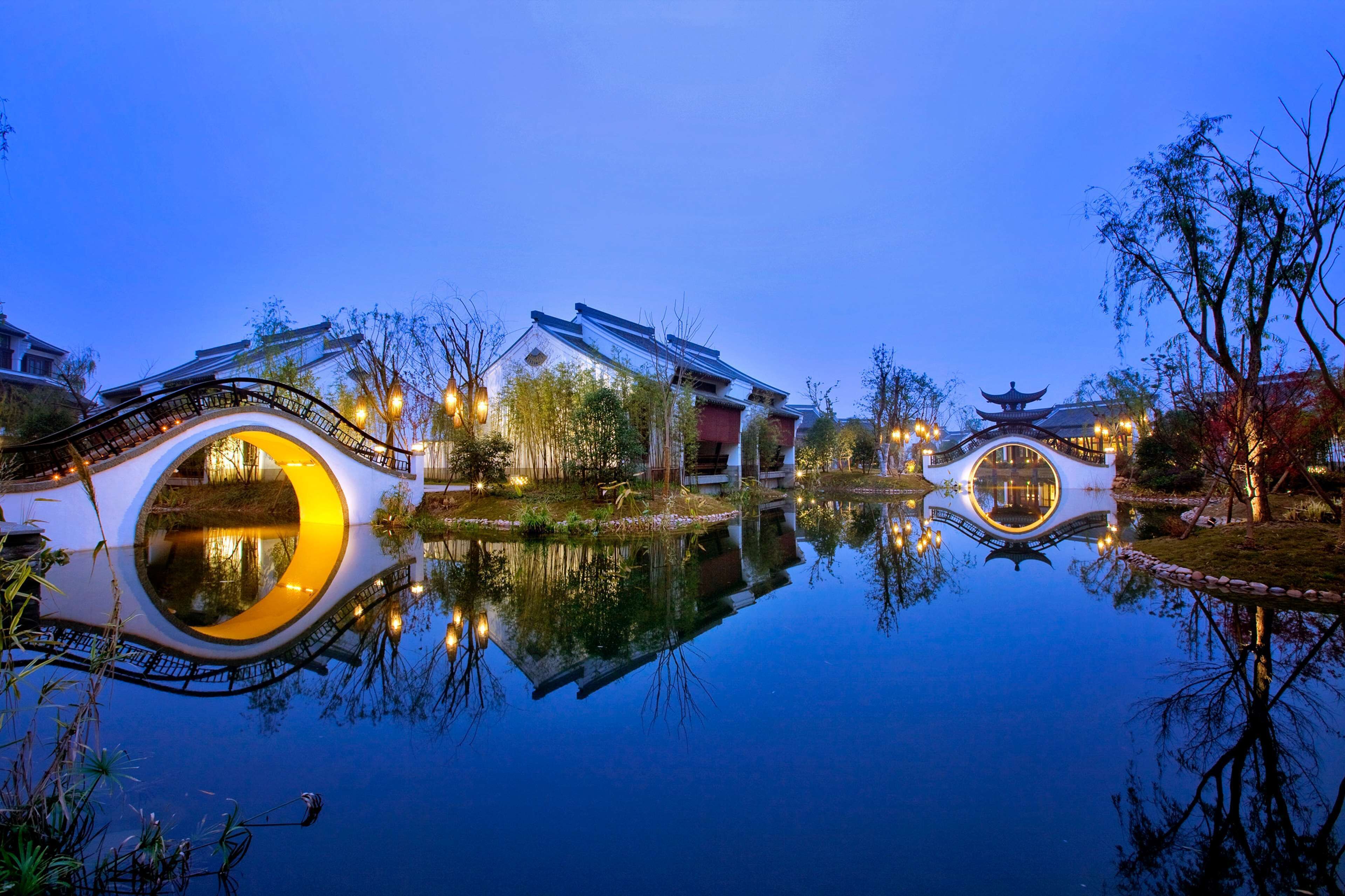 A serene lakeside setting with traditional architecture, including a bridge and pagoda-like structures, reflecting on the water.