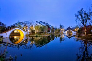 Un cadre paisible au bord d’un lac, avec une architecture traditionnelle comprenant un pont et des constructions ressemblant à des pagodes, dont le reflet se mire dans l’eau.