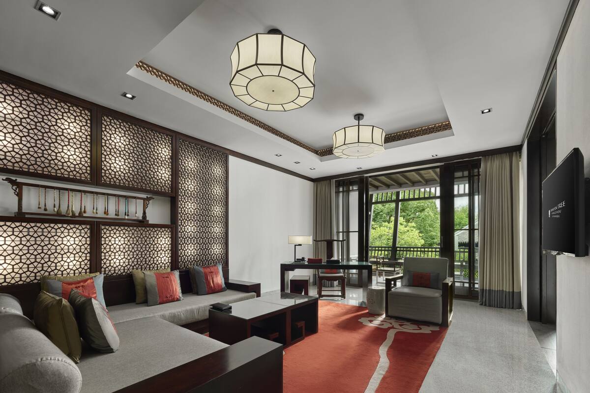 A modern living room with a sofa, coffee table, and TV. There is a decorative wall and a large window with a view of greenery.