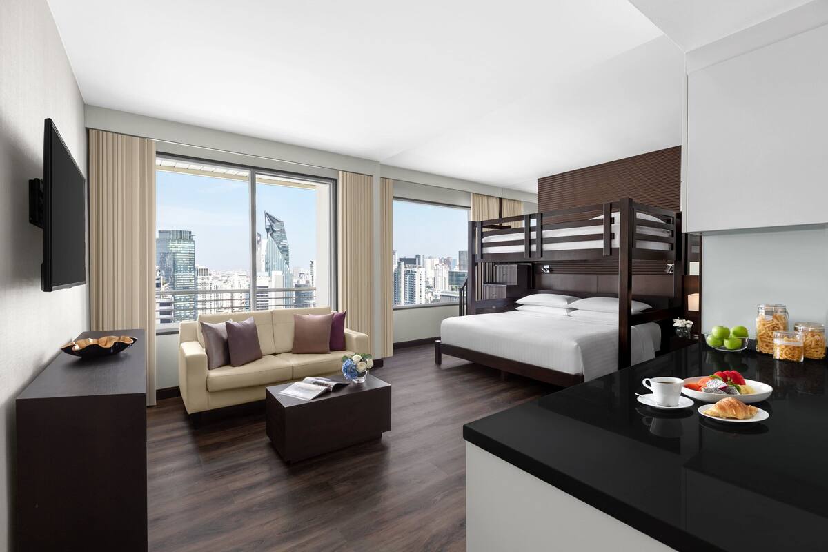 A modern hotel room with a city view, a bunk bed, a sofa, a coffee table, and breakfast items on the counter.