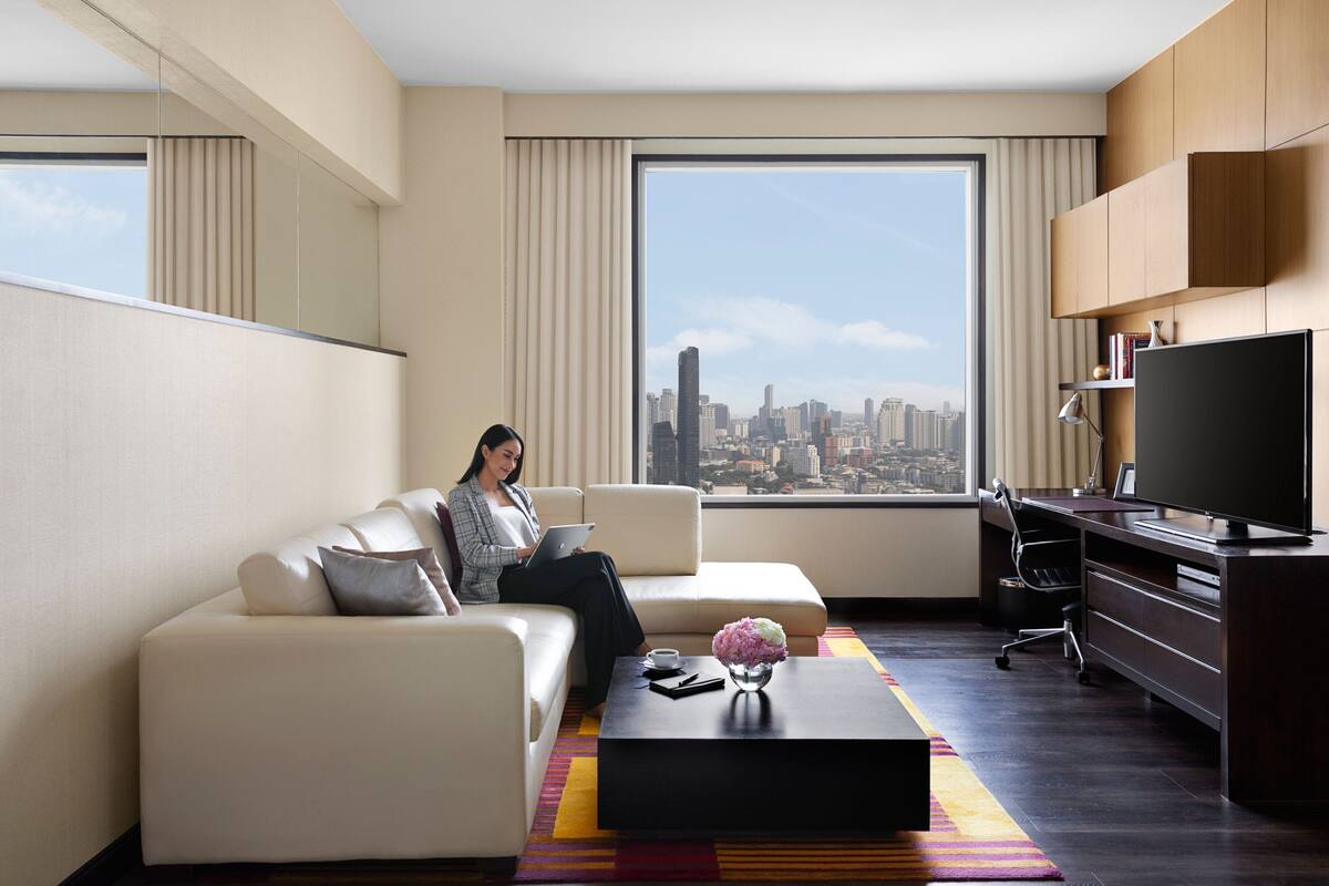 A modern hotel room with a large window offering a city view, a sofa, a desk, and a flat-screen TV.