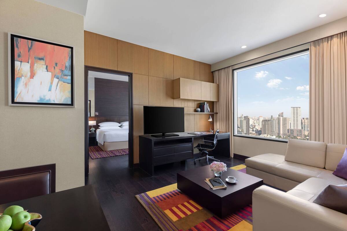 A modern hotel room with a large window, a flat-screen TV, a sofa, a desk, and a bed.