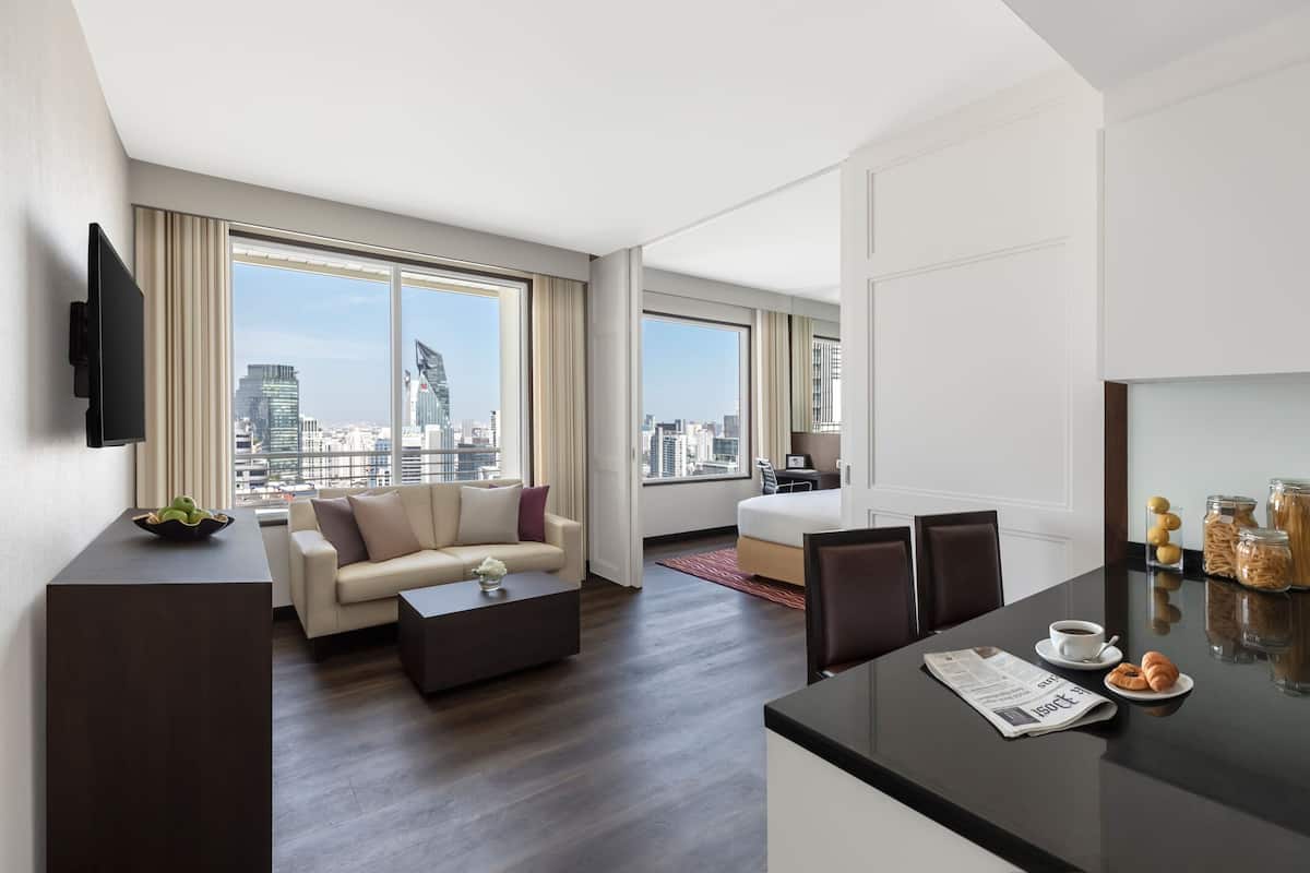A modern hotel room with a city view, a sofa, a coffee table, a dining area with a breakfast setup, and a flat-screen TV mounted on the wall.